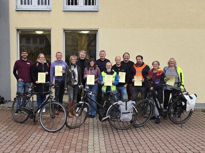 Bild vergr&ouml;&szlig;ern: Eine Gruppe von Frauen und M�nnern steht vor einer Hauswand und h�lt Urkunden in den H�nden. Im Vordergrund stehen drei Fahrr�der.
