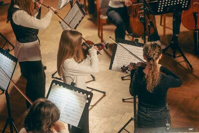Bild vergr&ouml;&szlig;ern: Kinder stehen auf einem Parkettboden und spielen auf Ihren Streichinstrumenten. Alle Kinder kehren der Kamera den R�cken zu.