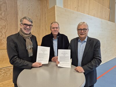 Bild vergrößern: Drei Mnner stehen an einem runden Tisch. Die beiden Mnner rechts und links halten jeweils ein Schriftstck in den Hnden und zeigen dies in die Kamera.