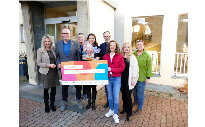 Bild vergr&ouml;&szlig;ern: Eine Gruppe von Frauen und M�nnern steht versetzt zueinander vor einer Hauswand. Zwei personen halten einen gro�formatigen Scheck in den H�nden. Alle l�cheln in die Kamera.