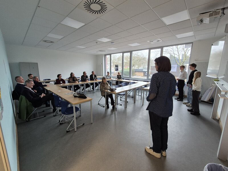 Das Bild zeigt einen Klassenraum. An den Tischen sind mehrere Personen zu sehen, die aufmerksam den Blick nach vorne auf drei Jugendliche und eine Lehrerin richten.