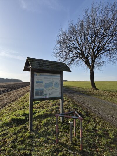 Bild vergr&ouml;&szlig;ern: Links von einem Feldweg steht eine �berdachte Hinweistafel aus Holz. Rechts im Hintergrund steht ein kahler Baum im Sonnenlicht.