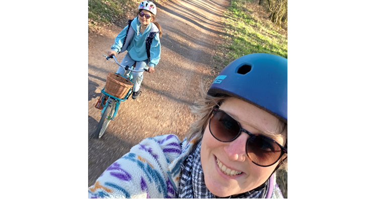Im Vordergrund ist das Gesicht einer Frau mit Sonnenbrille und Fahrradhelm zu sehen. Im Hintergrund f&auml;hrt ein Kind mit einem Rad auf einem Feldweg der Kamera entgegen.