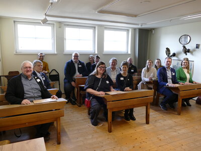 Bild vergr&ouml;&szlig;ern: Eine Gruppe von Frauen und M�nnern sitzt auf alten Schulb�nken in einem Raum mit Holzfu�boden.