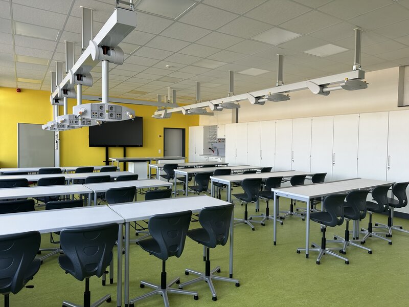 Das Foto zeigt einen Raum mit gelben W&auml;nden und wei&szlig;en Tischreihen, an dennen schwarze St&uuml;hle stehen.