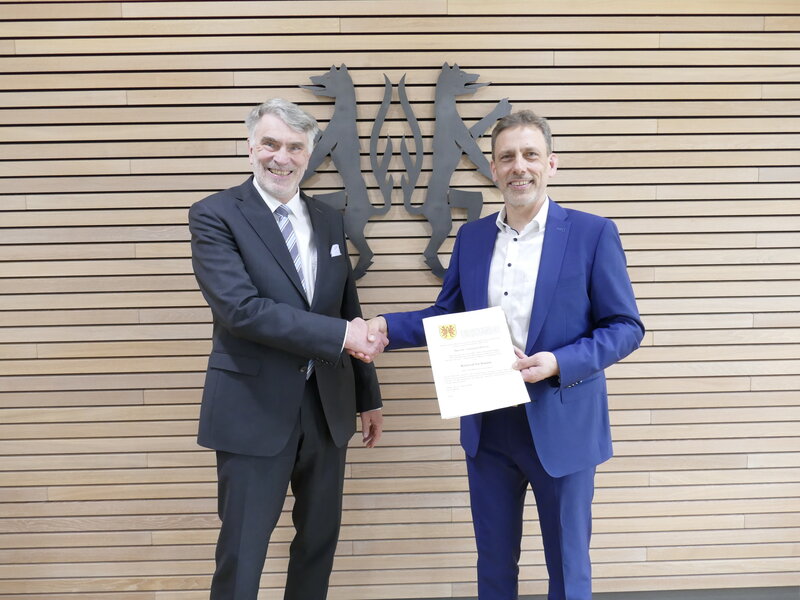 Zwei M&auml;nner stehen vor einer vert&auml;felten Wand, die das Kreiswappen in schwarz zeigt. Sie geben sich die Hand und l&auml;cheln in die Kamera.