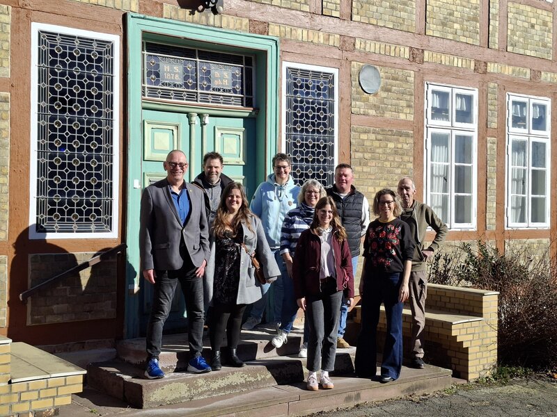 Eine Gruppe von Frauen und M&auml;nnern steht versetzt zueinander vor einer Haust&uuml;r.
