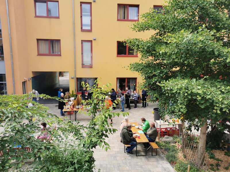 In einem Innhof, umgeben von Geb&auml;uden, stehen Bierzeltgarnituren, auf denen Menschen sitzen. Rund herum ist alles begr&uuml;nt.