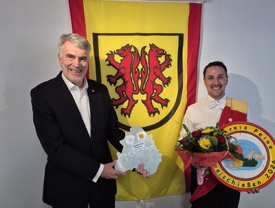 Bild vergr&ouml;&szlig;ern: Zwei M�nner stehen vor einer Wand mit einem Banner. Der Mann links h�lt eine Wanderplakette in den H�nden. Der Mann rechts tr�gt einen Blumenstrau� und eine K�nigsscheibe.