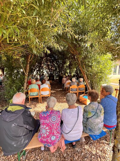 Bild vergr&ouml;&szlig;ern: Mehrere Gruppen von Personen sitzen auf Holzb�nken unter Weiden�sten.