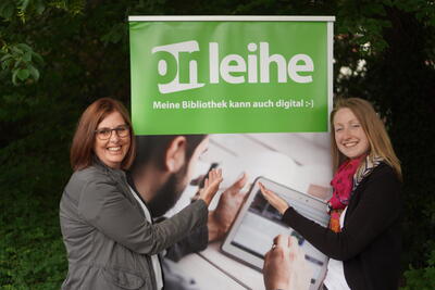 Bild vergrößern: Zwei Frauen halten gemeinsam ein Plakat in den Hnden und blicken lchelnd in die Kamera.
