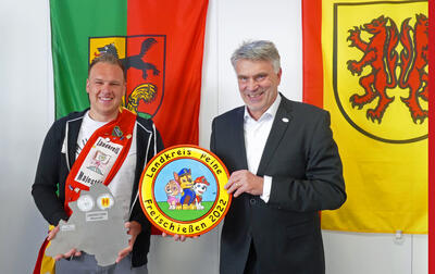 Bild vergrößern: Zwei Mnner stehen vor einer weien Wand, an der zwei Fahnen hngen. Der linke Mann trgt eine Schrpe, der rechte Mann hlt eine Schtzenscheibe in den Hnden.